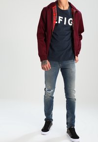 Chaqueta marrón con forro a cuadros sobre una camiseta azul marino con texto blanco, combinada con jeans cortos de mezclilla azul claro y zapatillas negras.