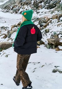 Osoba v zelené čepici a šále, černé bundě s nášivkou červeného jablka a kalhotách s leopardím vzorem, kráčející po zasněženém kamenitém terénu.