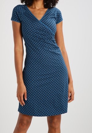 Kobieta ubrana w dopasowaną, krótkim rękawem, sięgającą do kolan niebieską sukienkę z niewielkim geometrycznym wzorem i dekoltem w stylu kopertowym.