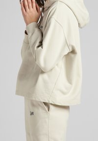 Personne portant un sweat à capuche Lee couleur crème et un pantalon assorti, vue de profil avec les mains proches du visage sur un fond neutre.