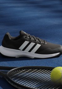 Μαύρο και άσπρο παπούτσι τρεξίματος Adidas δίπλα σε ρακέτα τένις και φωτεινή κίτρινη μπάλα τένις σε μπλε επιφάνεια γηπέδου.