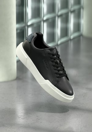 Black leather sneaker with white sole, black laces, and embossed branding, shown floating above a gray floor with glass block wall background.