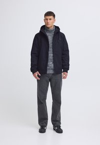 Black jacket with a hood, grey patterned sweater, grey jeans, and black shoes. The jacket features a zipper and side pockets.