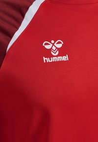 Rotes Sportshirt aus strukturiertem Stoff mit kontrastierenden dunkelroten Schultern und einem weißen Logo "hummel" auf der linken Seite.