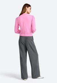 Femme de dos, portant un haut rose à manches longues, un pantalon large gris, des chaussures blanches et des boucles d'oreilles en argent.