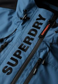 Niebieska kurtka z czarnym napisem "SUPERDRY", z zamkami, regulowanymi sznurkami oraz gładką, teksturowaną tkaniną. Akcenty w kontrastującym czarnym kolorze.