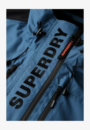Blå jakke med sort "SUPERDRY" tekst, der har lynlåse, justerbare snore og et glat, tekstureret stof. Accenter i kontrasterende sort.