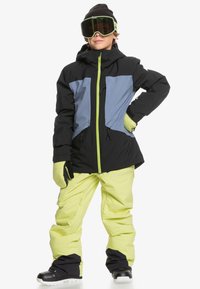 Completo da sci con giacca nera e azzurro chiaro con dettagli verde neon, abbinata a pantaloni gialli brillanti e stivali neri. Inclusi guanti.