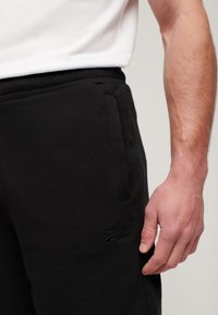 Pantaloni della tuta neri realizzati in morbido materiale, con una vita elastica, tasche laterali e un logo in rilievo discreto sulla coscia.