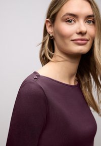 Mladá žena se světle hnědými vlasy, která má na sobě švestkový top s dlouhým rukávem s knoflíky na ramenou a malé zlaté kruhové náušnice, na jednobarevném pozadí.