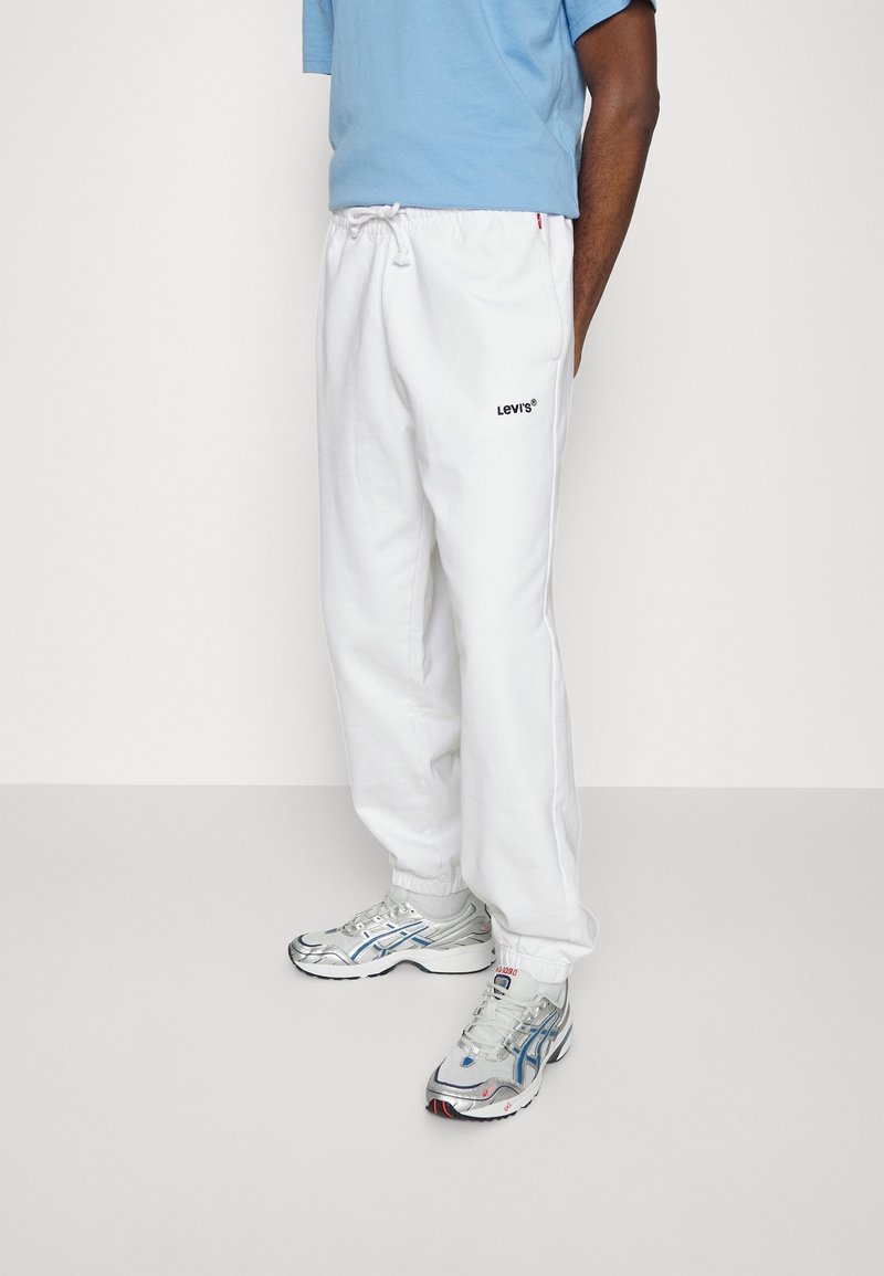 Homme portant un pantalon de jogging blanc Levi's avec des poignets élastiques, un t-shirt bleu clair et des baskets blanches et argentées, debout devant un fond uni.