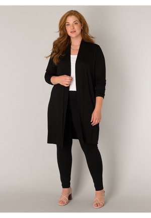 Vrouw met lang rood haar, gekleed in een zwarte openvallende cardigan, wit shirt, zwarte legging en beige sandalen met hak, staand tegen een grijze achtergrond.