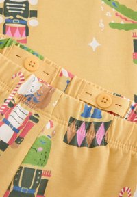 Tissu jaune avec des motifs festifs imprimés ; comprend des personnages de casse-noisette et des cannes en sucre. Présente deux boutons orange à la taille.