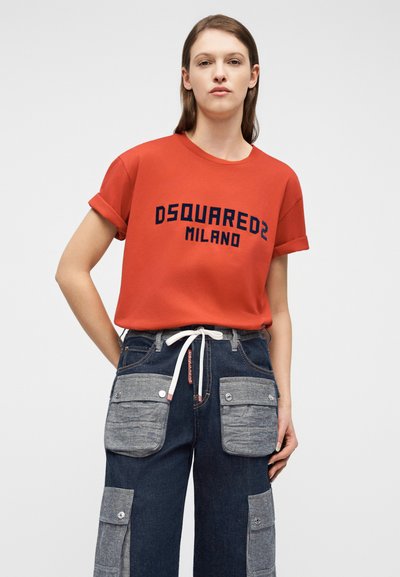 Νεαρό άτομο με μακριά μαλλιά φοράει πορτοκαλί μπλουζάκι Dsquared2 Milano και σκουρόχρωμο μπλε τζιν με μεγάλες γκρι τσέπες-μπαλώματα.