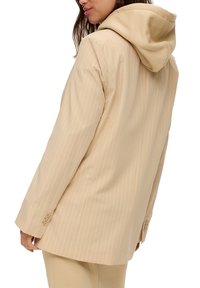Žena oděná v béžovém pruhovaném blejzru s připevněnou hnědou kapucí, zobrazena zezadu, s dlouhými hnědými vlasy a nosním kroužkem.