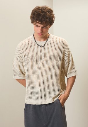 Junger Mann mit lockigem Haar, der einen weißen kurzärmeligen Grobstrickpullover und dunkle Hosen trägt, schaut nach unten, mit einer Hand in der Tasche.