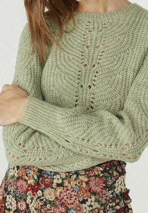 Femme vêtue d'un pull tricoté vert clair avec des motifs texturés croisés sur une jupe fleurie multicolore.