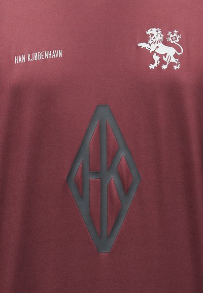 Maroontoonsest kangast särk, millel on musta kummist logo teemantkujul ja valge tekst "HAN KJØBENHAVN" koos lõvitempliga üleval.