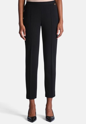 Pantalon noir slim fit longueur cheville avec pli sur le devant, porté avec des talons hauts noirs à bout pointu sur une personne debout.