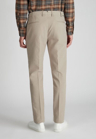 Uomo che indossa pantaloni chinos beige, sneakers bianche e una camicia a maniche lunghe a quadri marroni, in piedi davanti a uno sfondo grigio chiaro uniforme.