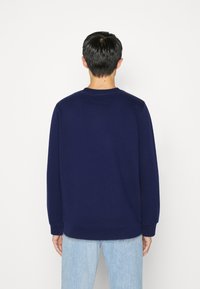Uomo di spalle con indosso una felpa lungo scuro blu semplice e jeans chiari blu su uno sfondo bianco.