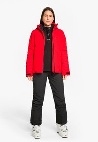 Red insulated ski jacket with a high collar, paired with a black sweater and black ski pants. White ski boots with blue accents.