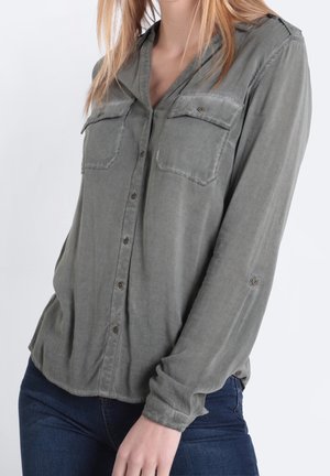 Frau trägt ein verblasstes grünes Hemd mit Knöpfen und zwei Brusttaschen sowie blaue Jeans, posiert mit einer Hand entspannt an ihrer Seite.