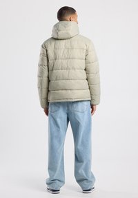 Chaqueta tipo puffer en verde claro, con diseño acolchado y capucha. Combinada con jeans holgados de color azul. Estilo casual de streetwear.