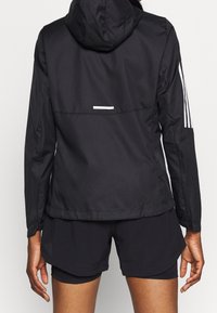 Schwarze, leichte Jacke mit Kapuze, aus strukturierter Stoffqualität, weißen reflektierenden Akzenten und drei weißen Streifen an den Ärmeln.