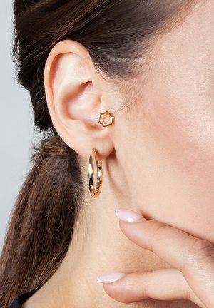 Złote kolczyki w formie obręczy z kręconym wzorem oraz sześciokątne kolczyki wkręty. Oba mają błyszczące metaliczne wykończenie. Tło w odcieniu skóry.