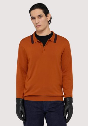 Polo a maniche lunghe color ambra con dettagli del colletto in viola scuro, dotato di due bottoni al collo. Il modello indossa guanti neri e pantaloni blu navy.