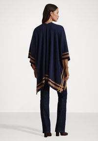 Donna in piedi, che indossa un poncho blu navy con strisce marroni sopra maniche marroni, jeans scuri e stivali con tacco marroni, di spalle.