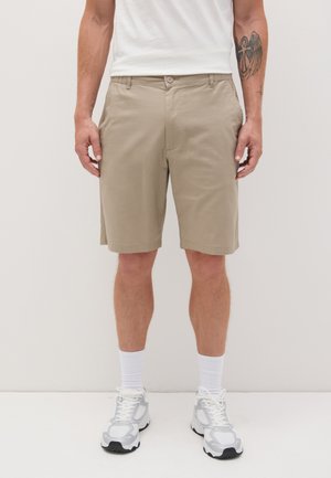 Beige cotton shorts with front pockets and a button closure. Worn with a plain white t-shirt and gray sneakers. Simple, casual design.
