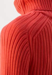 Gros plan sur un tissu en maille côtelée orange vif, mettant en évidence la couture de l'épaule et la texture du pull à col roulé.