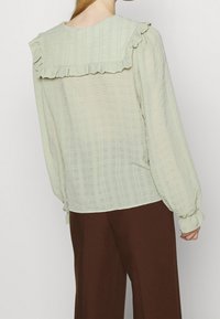 Light green, textured blouse with ruffled collar and balloon sleeves, paired with brown wide-leg pants. Subtle striped pattern visible.