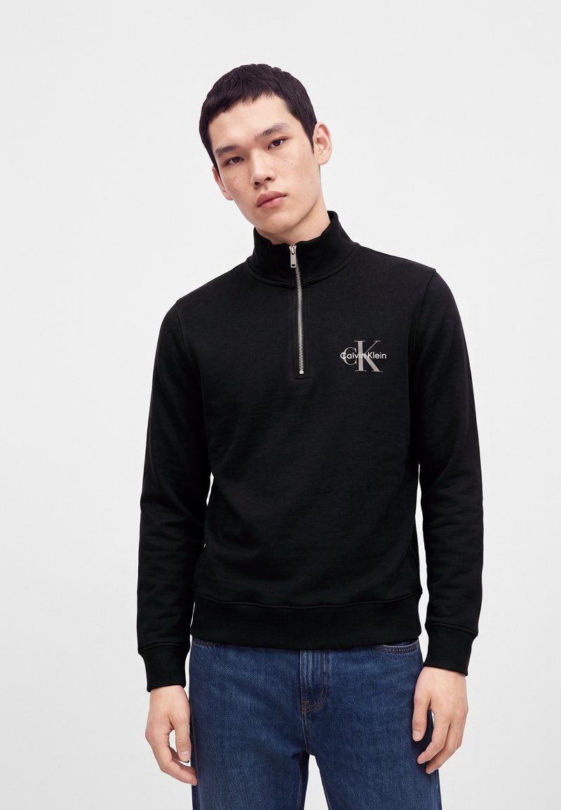 Jeune homme portant un sweat-shirt noir Calvin Klein à fermeture éclair quart de fermeture et un jean bleu, debout devant un fond clair uni.