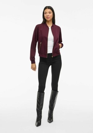 Femme portant une veste zippée bordeaux, une chemise blanche, un pantalon noir et des bottes noires jusqu'aux genoux, se tenant devant un fond blanc.