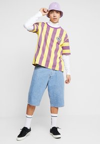 Mehrfarbig gestreiftes T-Shirt in Lila und Gelb, übereinander getragen mit einem weißen Langarmshirt. Denim-Shorts und schwarze Sneakers runden das Outfit ab.