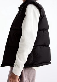 Černá prošívaná vesta s vysokým límcem, zapínáním na zip a bočními kapsami, vrstvená přes bílou košili s dlouhým rukávem, s jemnou texturou.