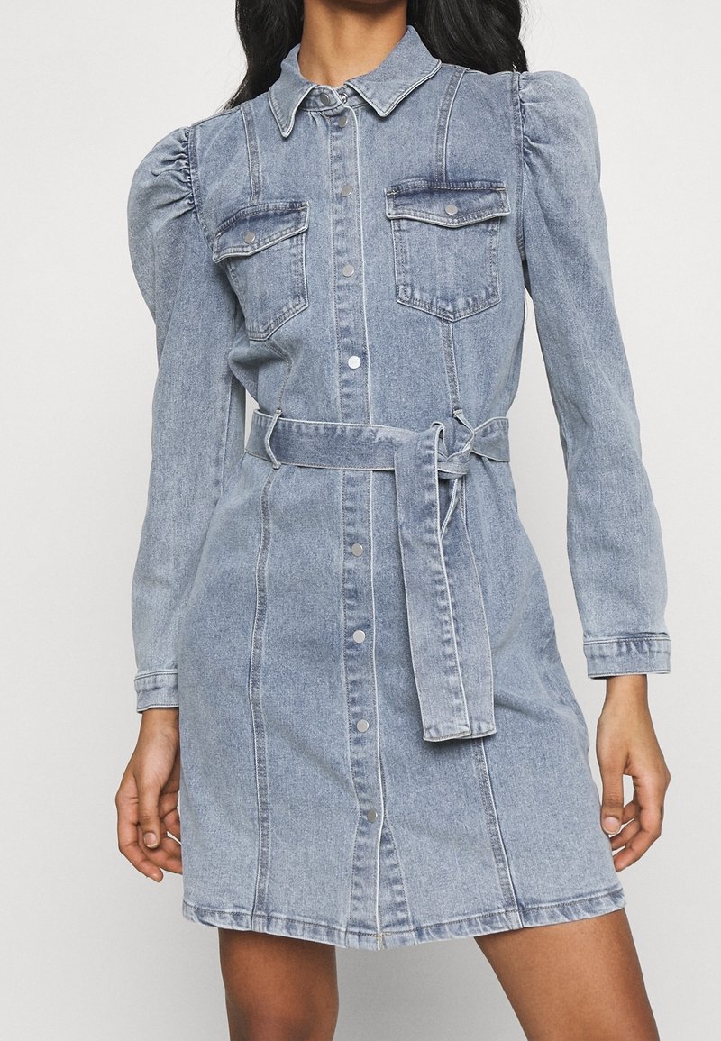 Femme portant une robe en denim bleu clair avec des manches longues bouffantes, des poches avant, une fermeture à boutons et une ceinture nouée à la taille.