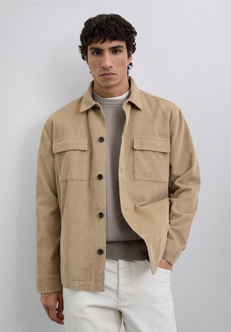 Joven vestido con una chaqueta de pana beige sobre un suéter gris y pantalones blancos, de pie con una mano en el bolsillo frente a un fondo sencillo.