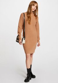 Gebreide jurk in een warme beige kleur, met een hoge halslijn, lange mouwen en een getailleerde pasvorm. Gecombineerd met zwarte enkellaarzen.