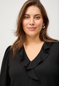 Black blouse with textured fabric, featuring a V-neckline and ruffled front detail. Model has medium-length brown hair and wears hoop earrings.