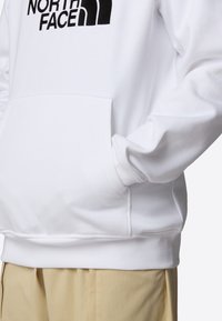 Sweat à capuche blanc avec le logo noir "The North Face", porté avec un pantalon beige, mains dans la poche avant côté droit.