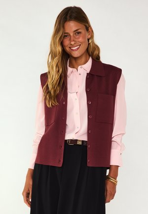 Donna sorridente con lunghi capelli biondi che indossa un gilet bordeaux abbottonato sopra una camicia rosa chiaro e una gonna nera, accessoriata con braccialetti.