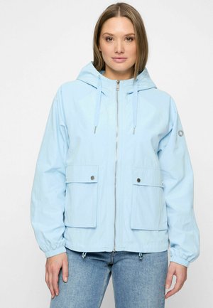 Vrouw draagt lichtblauwe gewatteerde jas met capuchon, voorzakken en rits, gecombineerd met een blauwe spijkerbroek, tegen een effen witte achtergrond.