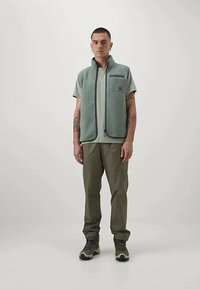 Homem com um colete de lã verde sobre uma t-shirt verde, calças oliva e botas de caminhada, de pé contra um fundo claro e simples.