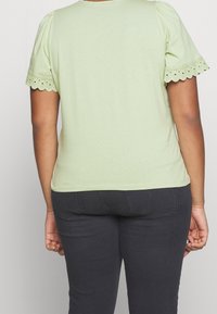 Top verde claro de manga corta con detalles de ganchillo a lo largo del dobladillo en forma de onda. Tejido suave y diseño ajustado. Combinado con jeans gris oscuro.