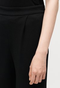 Gros plan de la main d'une personne reposant à côté d'un pantalon noir texturé avec une ceinture ajustée sur un fond clair.