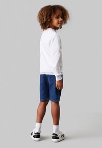 Felpa bianca e pantaloni shorts in denim blu indossati con calzini bianchi e sneakers nere, che mostrano un outfit casual e confortevole.